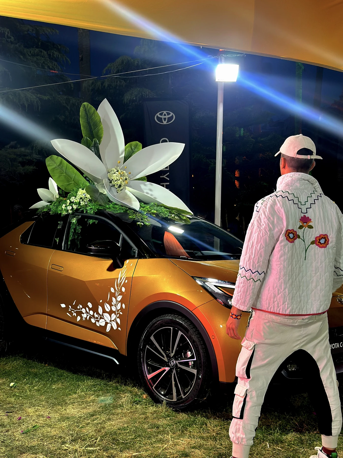 Tamer in a white jacket decorated with flowers beside the blossom-topped orange Toyota C-HR at night